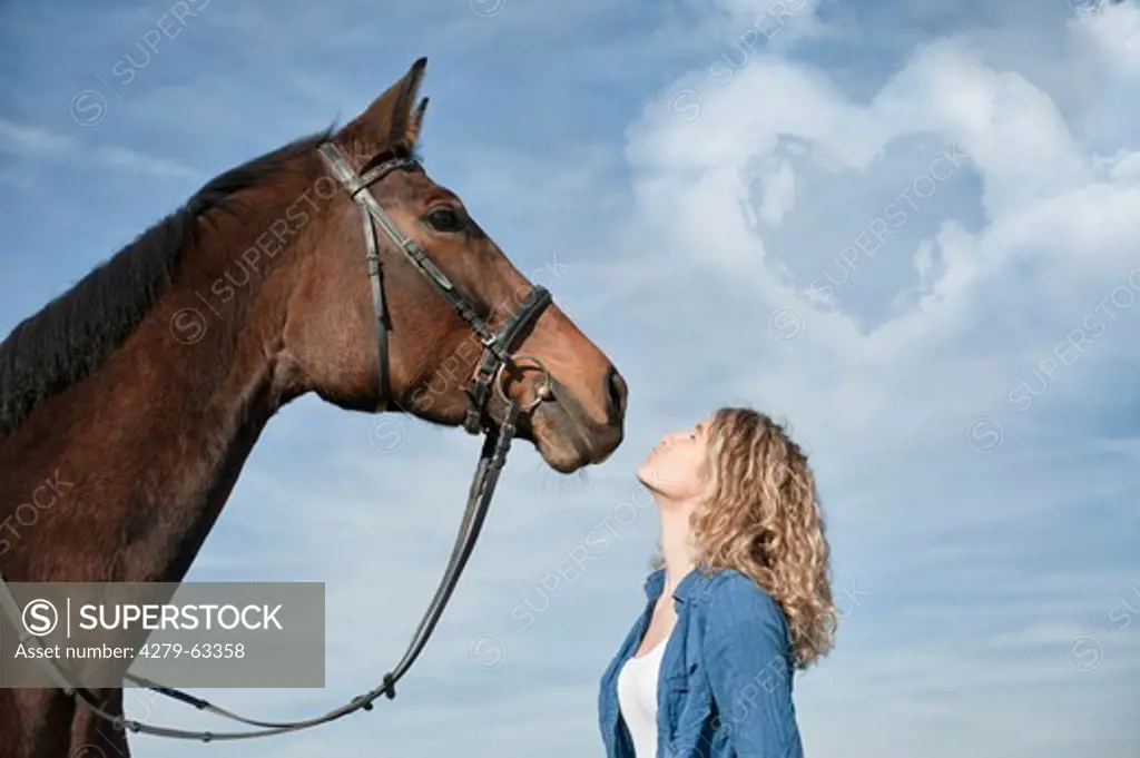 A young woman stands blowing a kiss while standing in front of a horse with heart-shaped cloud in the sky