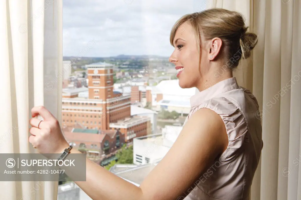 England, West Midlands, Birmingham. Woman in a hotel suite pulling back the curtains to look out over the city of Birmingham.