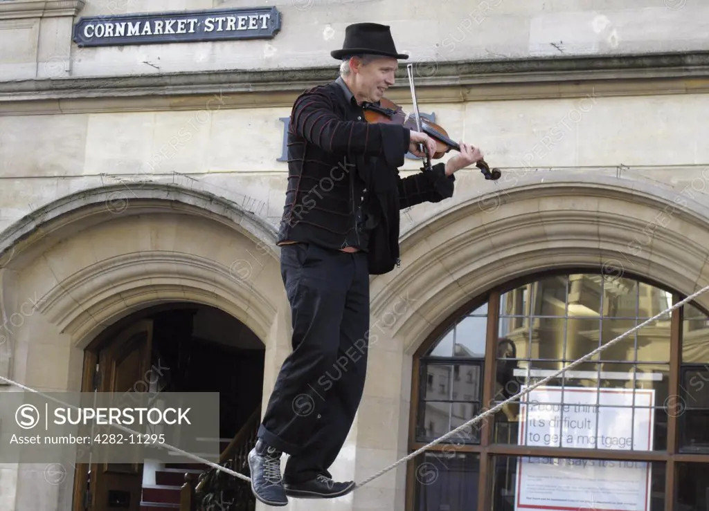 England, Oxfordshire, Oxford. A man playing the fiddle balances on a tightrope outside a bank premises.