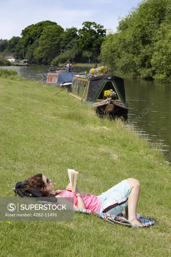 England, Oxfordshire, Abingdon. A young woman relaxing with a book on the river bank of the River Thames at Abingdon Bridge.