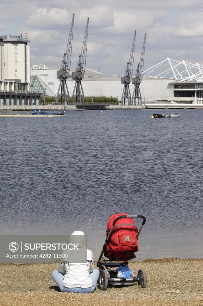 England, London, Docklands. A mother and child in a push-chair sitting on a bank of the River Thames looking towards the Royal Victoria Docks and the Excel Centre in London's Docklands.
