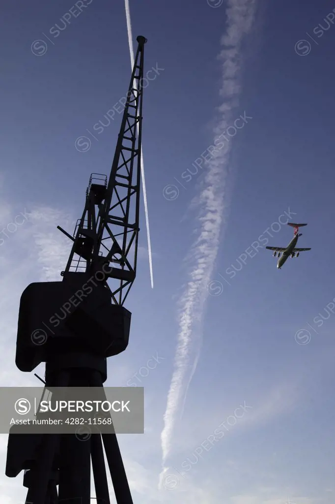 England, London, Royal Victoria Dock. A plane flying over the silhouette of a derelict crane at Royal Victoria Dock in the redeveloped Docklands area of London.