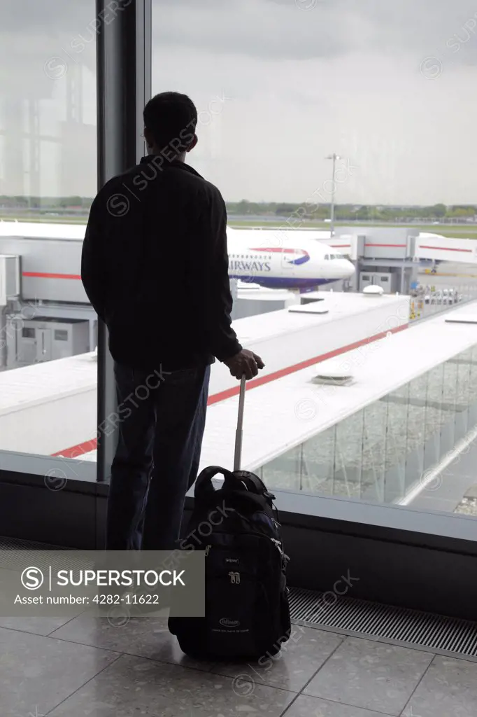 England, London, Heathrow. Silhouette of a passenger standing with his travel bag looking out of a window in the departure lounge of Heathrow Airport.