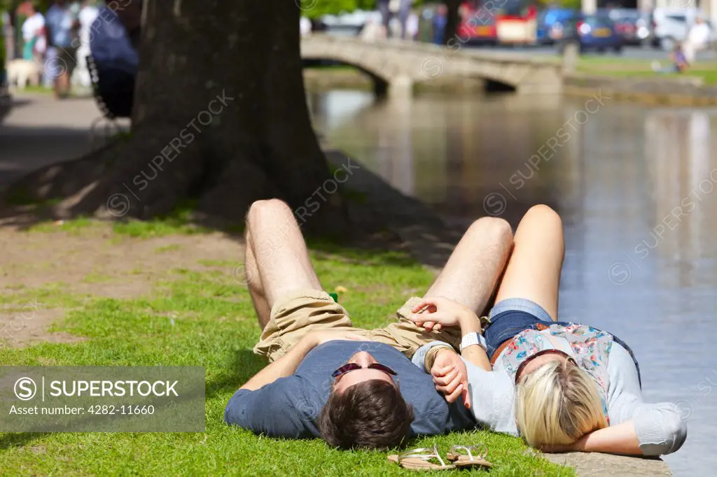 England, Gloucestershire, Bourton-on-the-Water. A couple relaxing on the riverbank of the River Windrush at Bourton-on-the-Water in the Cotswolds.