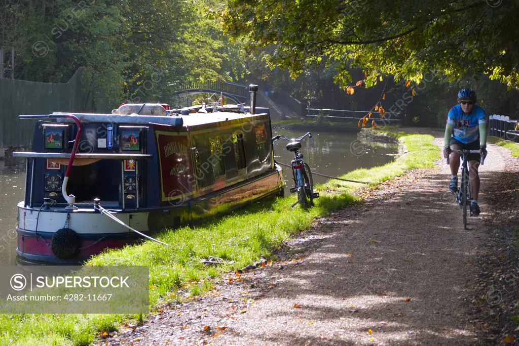 England, Oxfordshire, Oxford. A cyclist riding along the towpath past a houseboat on Oxford Canal in Autumn.
