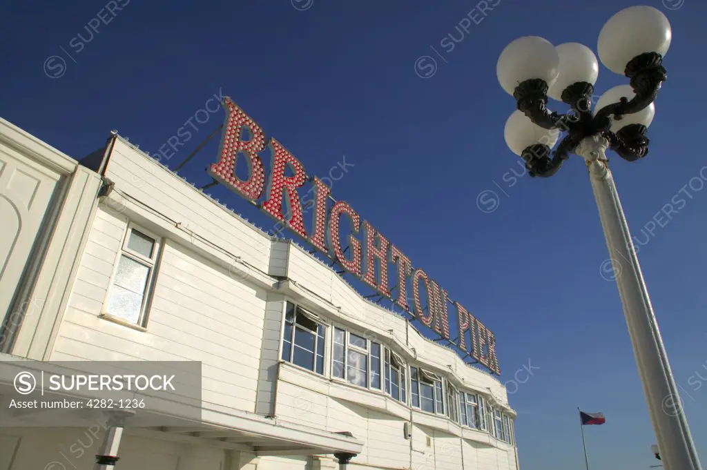 England, East Sussex, Brighton. An ornate lamppost opposite a Brighton Pier sign against a bright blue sky.