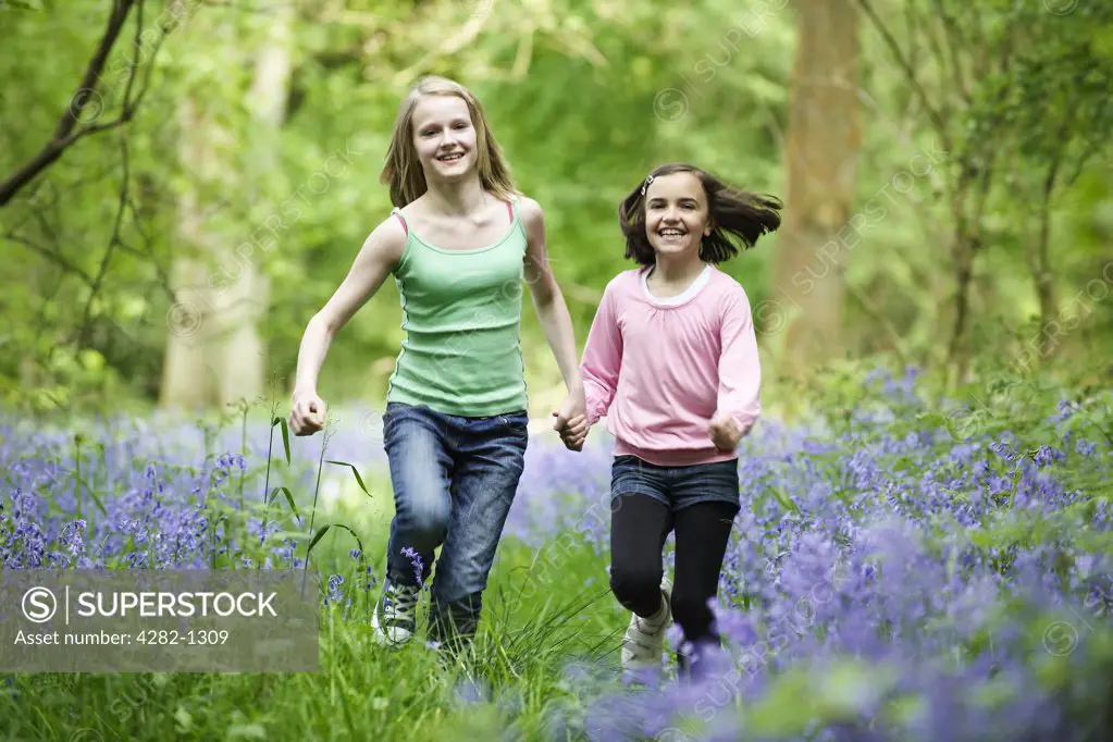 England, Buckinghamshire, Stokenchurch. Two young girls running hand in hand through a wood full of Bluebells.