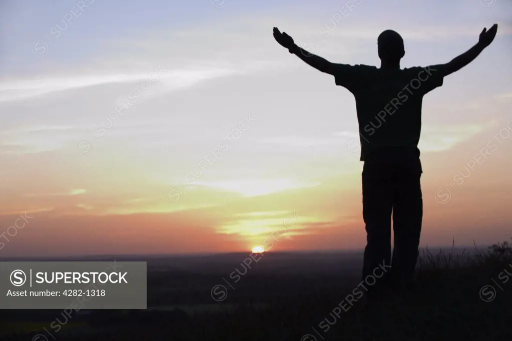 England, Buckinghamshire, Stokenchurch. The silhouette of a man standing on the Chiltern Hills with his arms outstretched.