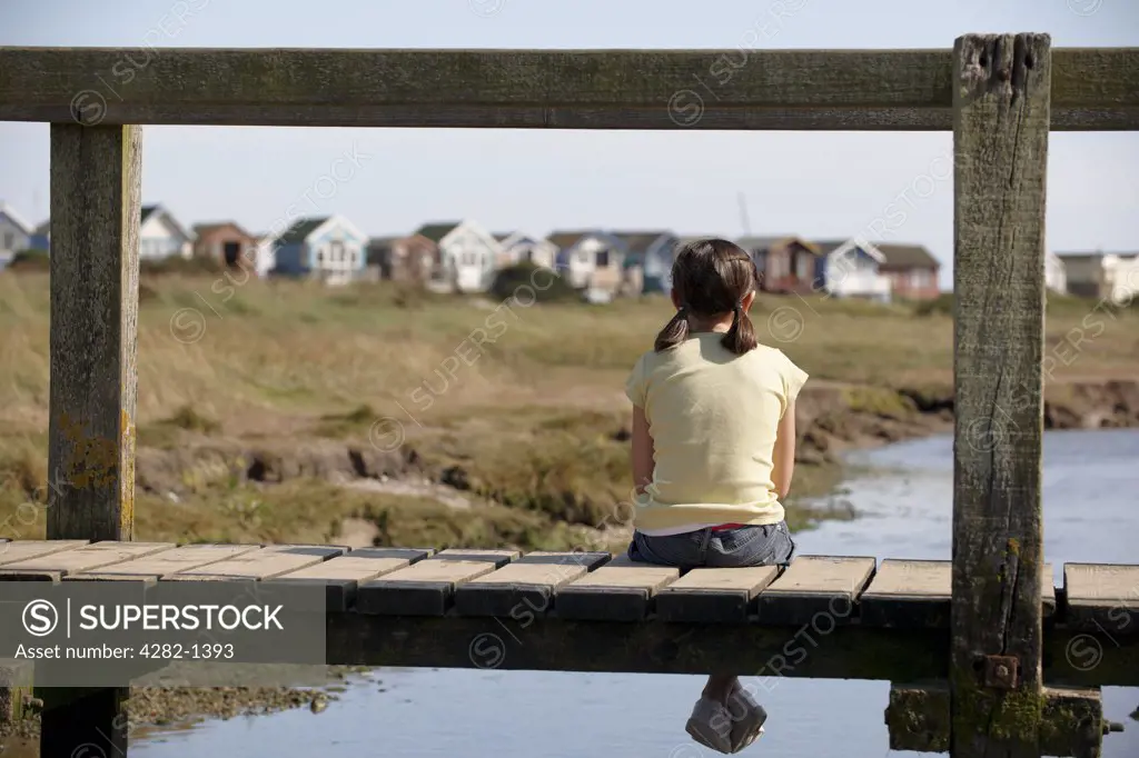 England, Dorset, Christchurch. A young girl sitting on a foot bridge looking towards beach huts in the distance at Christchurch in Dorset.
