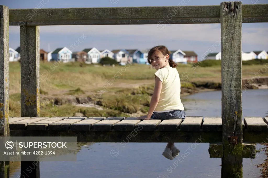England, Dorset, Christchurch. A young girl sitting on a foot bridge looking back over her shoulder with beach huts in the background.