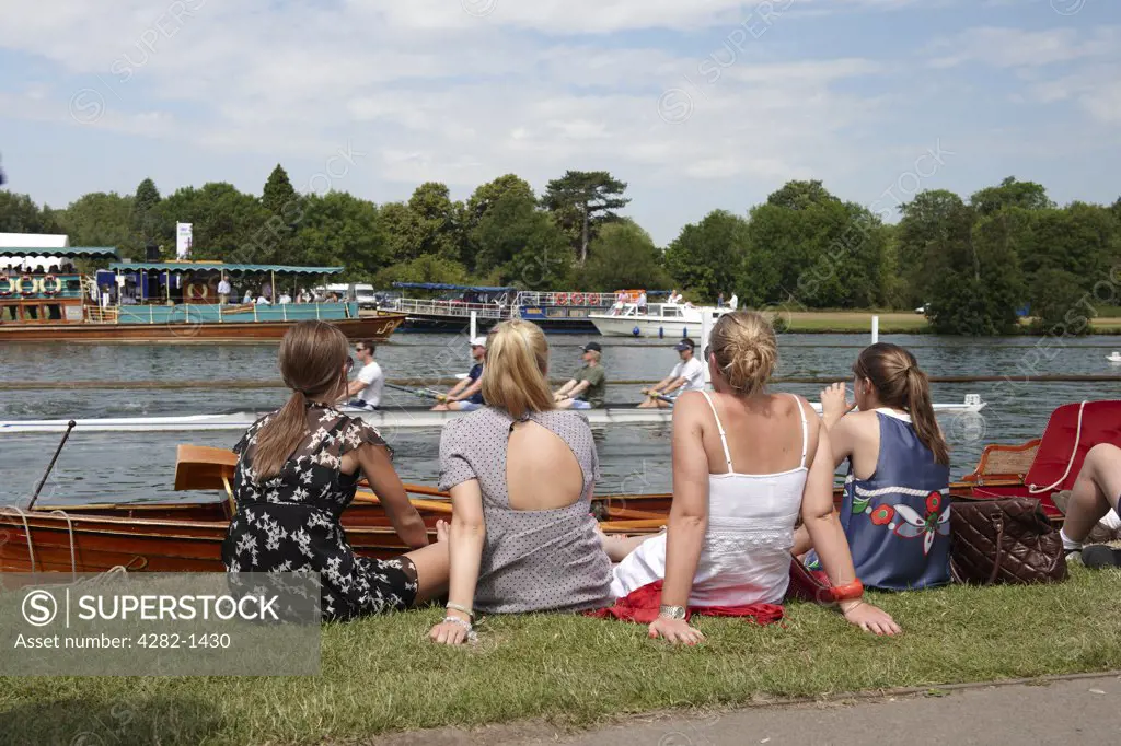 England, Oxfordshire, Henley-on-Thames. Four young women sitting on the riverbank enjoying the annual Henley Royal Regatta.