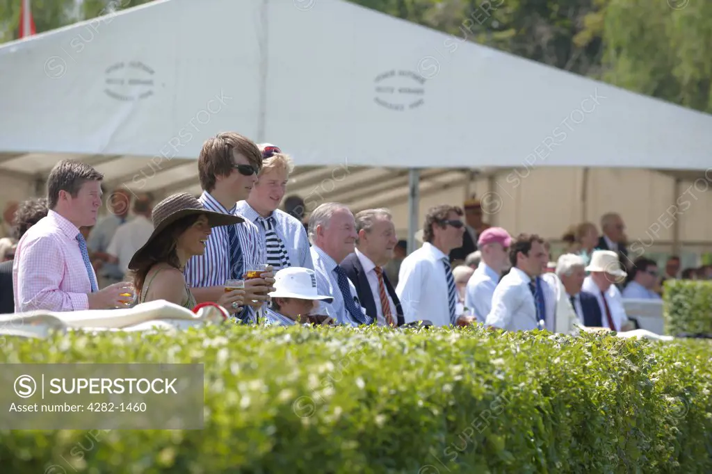 England, Oxfordshire, Henley-on-Thames. Spectators at the riverside by a hospitality marquee watching a race at the annual Henley Royal Regatta.