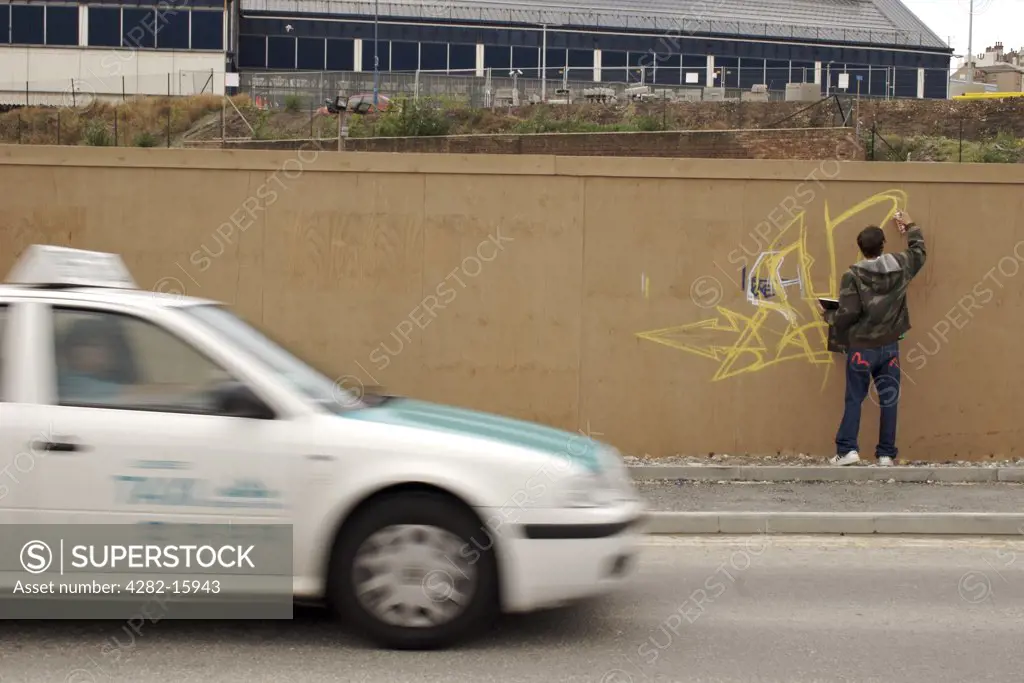 England, East Sussex, Brighton. A graffiti artist sketches as a taxi drives past. Graffiti artist sketches as taxi passes The word 'graffiti' is the plural of 'graffito' and originates from the Italian word 'graffiato' which means scratched.