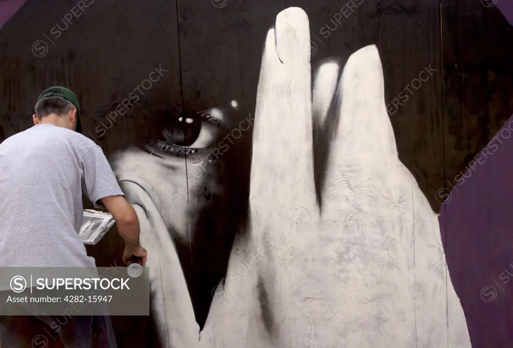 England, East Sussex, Brighton. Graffiti artist in front of portrait. The word 'graffiti' is the plural of 'graffito' and originates from the Italian word 'graffiato' which means scratched.