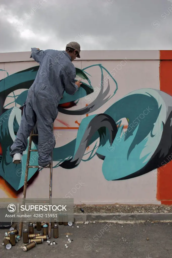 England, East Sussex, Brighton. Graffiti artist leans off a ladder to paint. The word 'graffiti' is the plural of 'graffito' and originates from the Italian word 'graffiato' which means scratched.