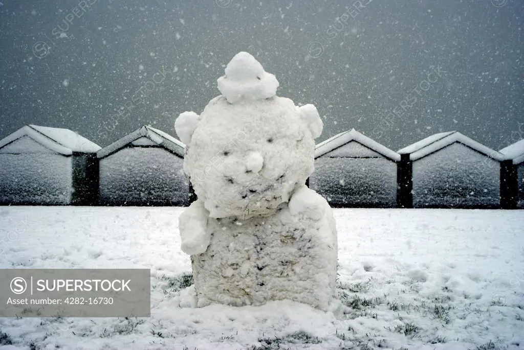 England, City of Brighton and Hove, Hove. A snowman on Hove Lawns with snow covered beach huts in the background.