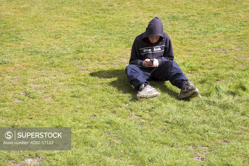 England, Tyne and Wear, Gateshead. A hooded teenager sitting on grass sending a text message from his mobile phone.