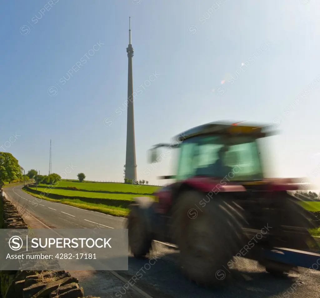 England, West Yorkshire, Emley Moor. A tractor travelling along a road on Emley Moor past the Emley Moor Tower, a television transmission tower which is the tallest free-standing building in the UK.