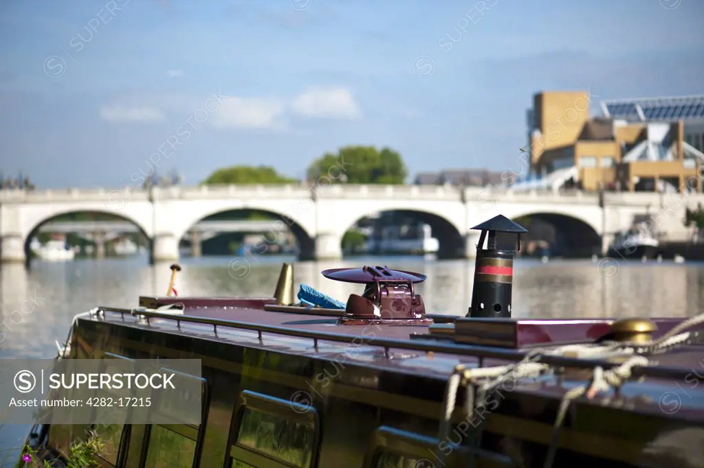 England, Surrey, KIngston Upon Thames. A canal barge moored on the River Thames with Kingston Bridge in the background.