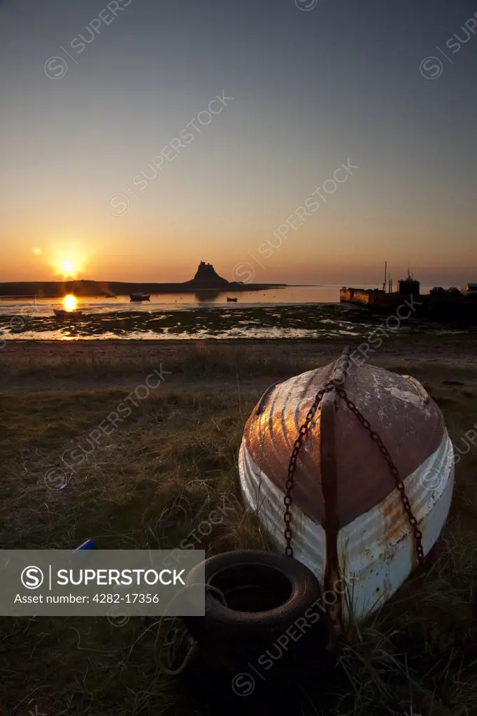England, Northumberland, Holy Island. A small upturned boat on the shore with Lindisfarne Castle silhouetted by dawn sunlight in the background.