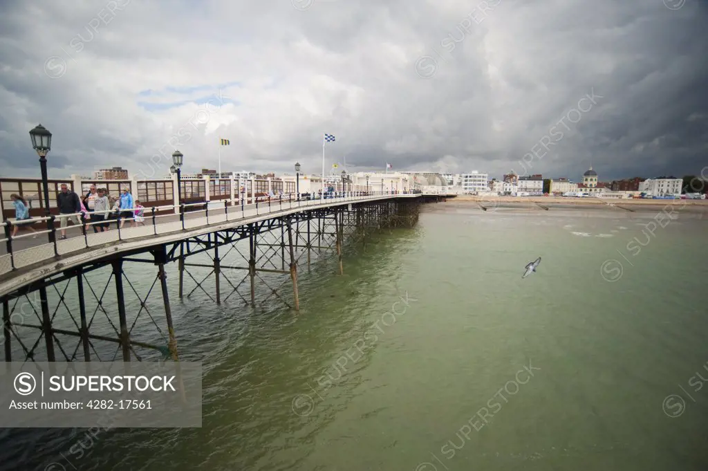 England, West Sussex, Worthing. Tourists walking along the Pier at Worthing with grey clouds over the town.