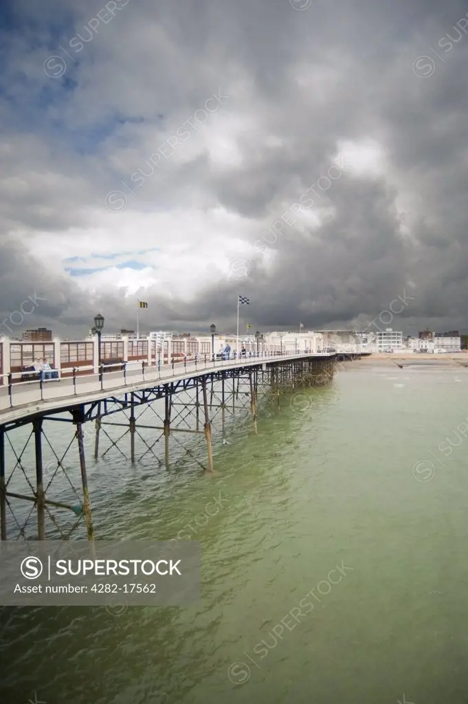 England, West Sussex, Worthing. Tourists walking along the Pier at Worthing with grey clouds over the town.