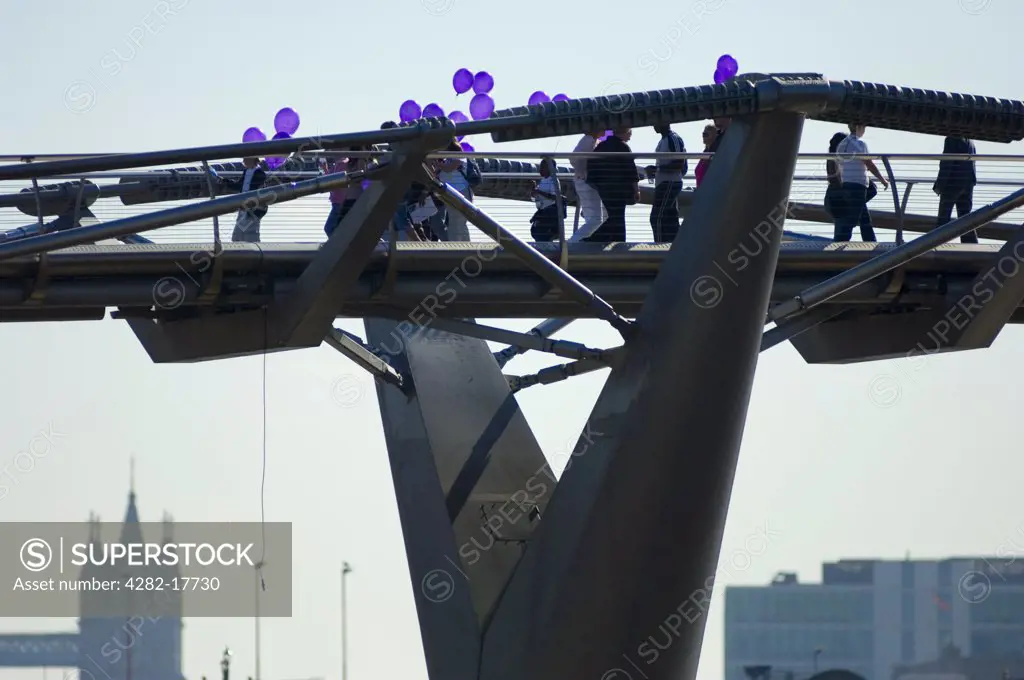 England, London, Millennium Bridge. Pedestrians holding purple balloons crossing the Millennium footbridge over the River Thames in central London with the Tower Bridge in the background.