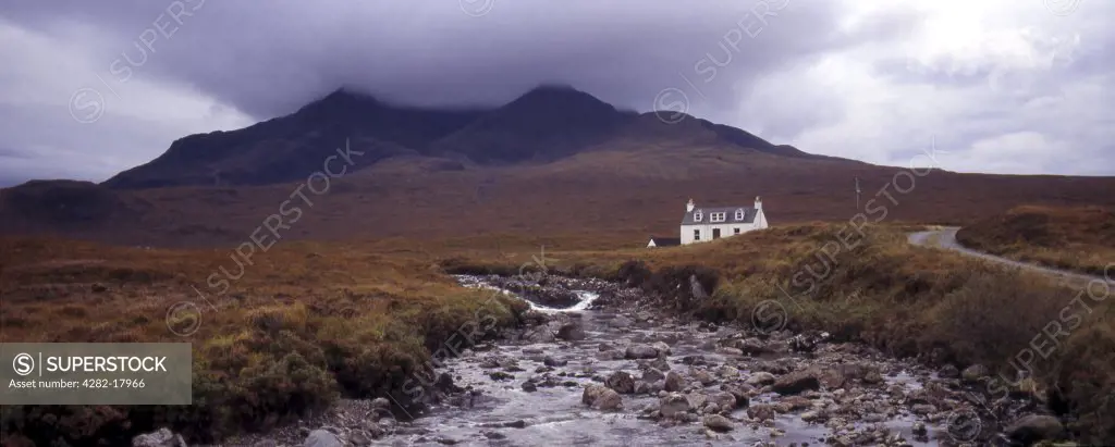 Scotland, Isle of Skye, Glen Sligachan. Panoramic view of cottage with river in foreground and cloud covered mountains in the background.