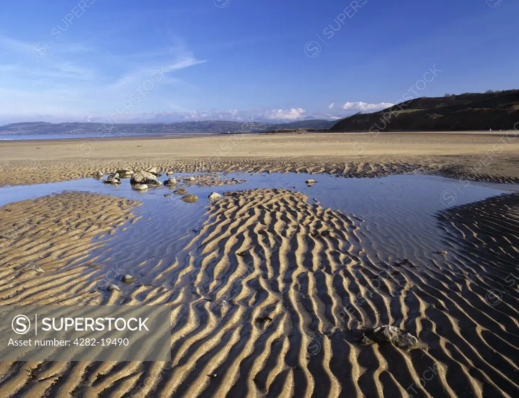 Wales, Anglesey, Benllech. Rippled sand on empty beach at low tide with low sun in late winter and snow on mountains of Snowdonia beyond.