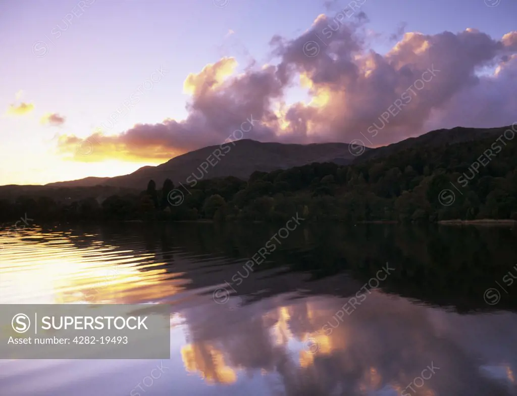 England, Cumbria, Coniston. Coniston Water at sunset with sun setting behind cloud reflected in calm water.