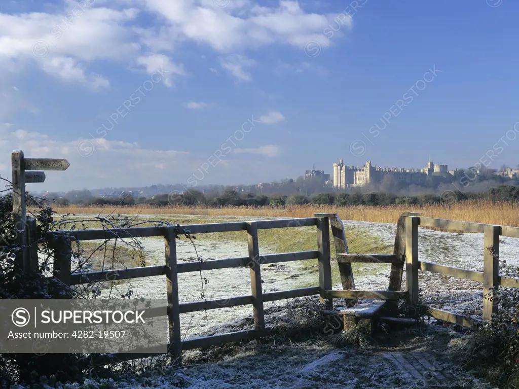 England, West Sussex, Arundel. Footpath and stile in frosty landscape on reed-lined embankment of River Arun with Arundel Castle beyond in winter.