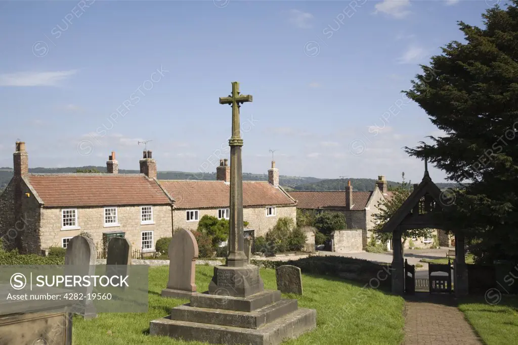 England, North Yorkshire, Coxwold. War memorial cross in church yard and traditional sand stone cottages in a village on the edge of North York Moors National Park.