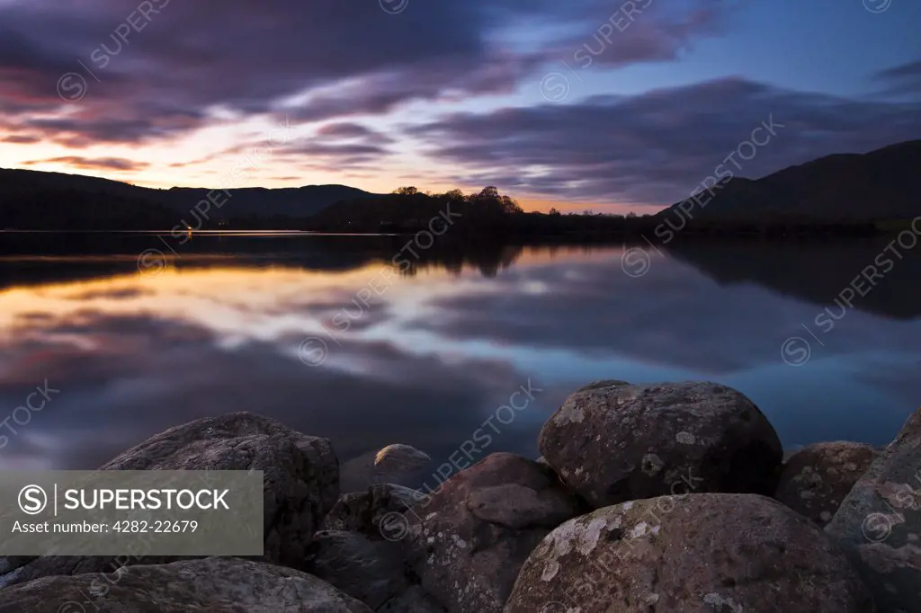 England, Cumbria, Keswick. A view over rounded rocks on the shore of Derwentwater at sunset.