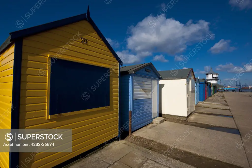 England, Essex, Harwich. Freshly painted beach huts on the beach at Harwich. Felixstowe Docks can be seen in the background.