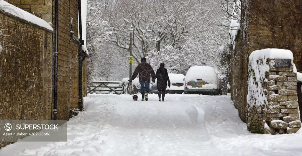 England, Gloucestershire, South Cerney. A couple walking a small dog through heavy snow in a Cotswold village.