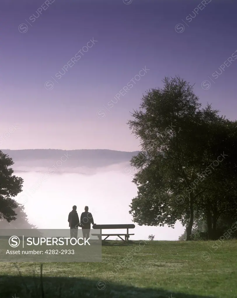 England, Surrey, Guildford. Couple stand by a bench on a misty October morning at Newlands Corner.