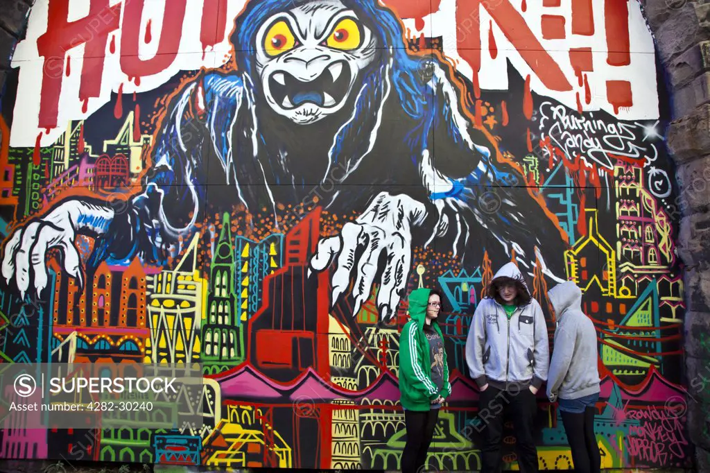 England, Tyne and Wear, Gateshead. Three teenagers wearing hooded tops in front of a large piece of street art depicting 'Horror'.