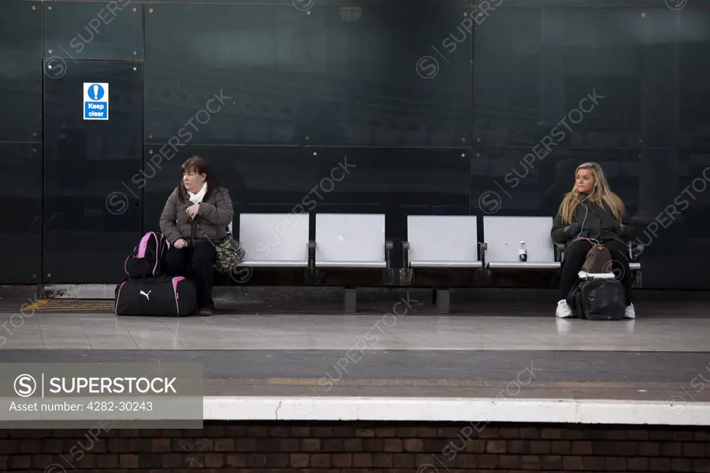 England, Tyne and Wear, Newcastle. Two women sitting at either end of a row of seats, waiting for a train on a platform at Newcastle Central Station.