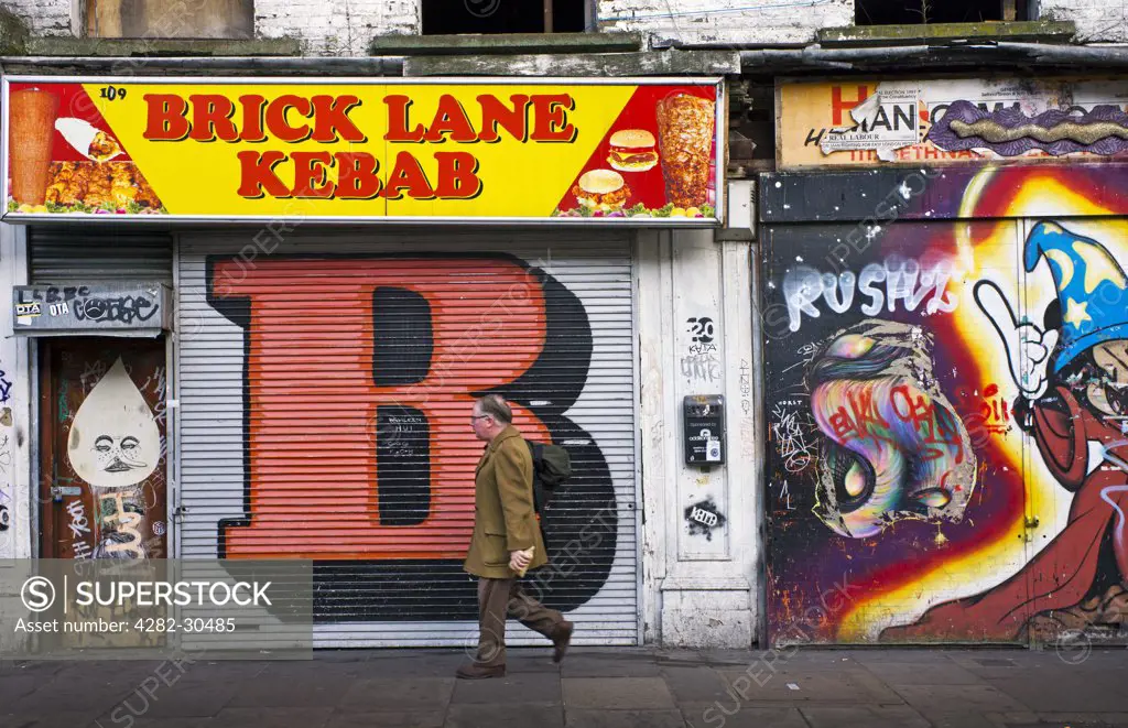 England, London, Shoreditch. A man walking to work past shopfronts covered in graffiti on Brick Lane in the East End of London.
