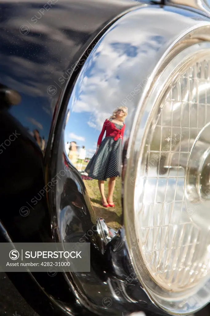 England, West Sussex, Goodwood. A girl in a black and white polka dot dress reflected in the chrome rim of a classic car's headlight at Goodwood Revival.