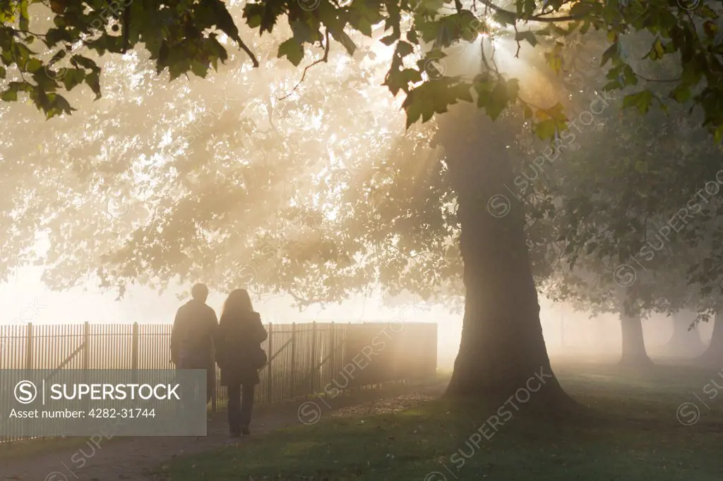 England, Oxfordshire, Christ Church. A couple walking on Christ Church Meadows on a misty autumn morning by the river Thames at Oxford.