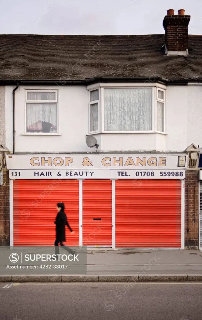 England, Essex, Rainham. A woman walking past a closed beauty salon with red shutters in Rainham.