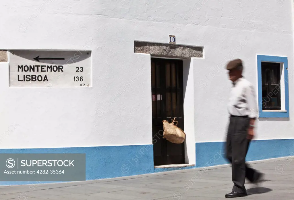 Portugal, Alentejo, Arraiolos. An elderly local man walks past a sign to Lisbon on the wall of a traditional blue-trimmed house.