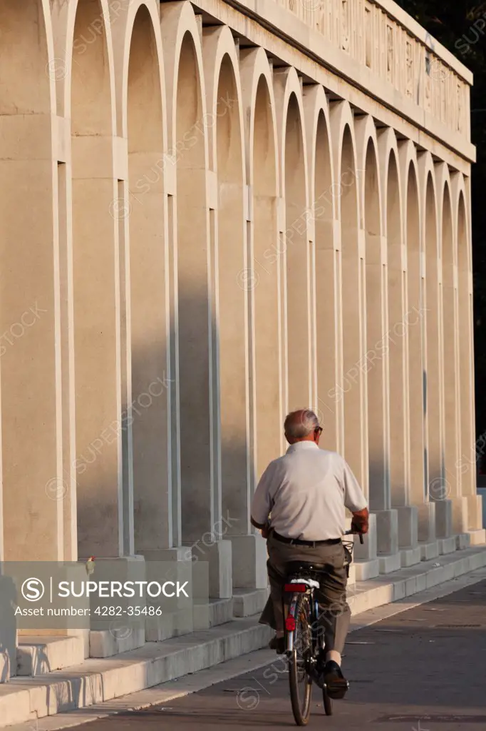 Italy, Emilia-Romagna, Tresigallo. An elderly cyclist passes by a Rationalist architecture arcade in Tresigallo.