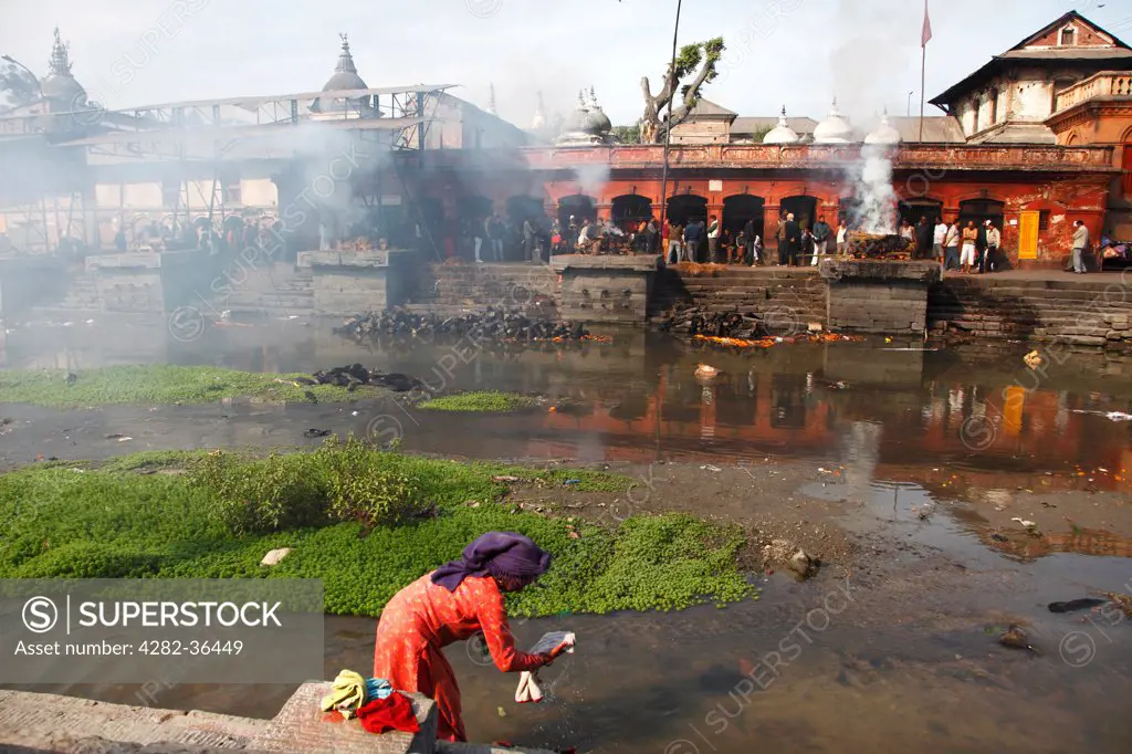 Nepal, Kathmandu, Pashpatinath. Cremation in Pashpatinath while a woman washes clothes on the Bagmatin riverbank.