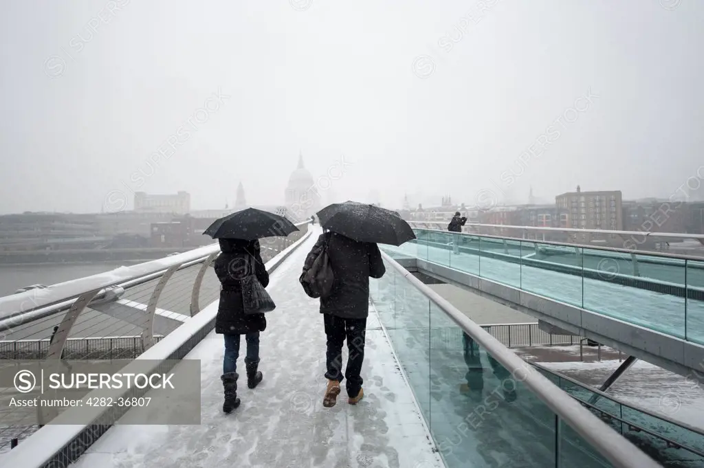 England, London, Southwark. Pedestrians crossing the Millennium Footbridge on a misty winter day sheltered from snowfall with umbrellas.
