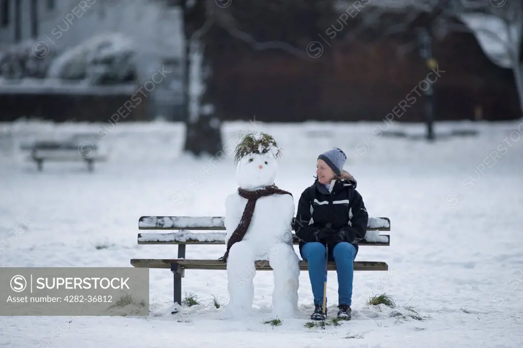 England, London, Wimbledon. Winter day with a woman walker sitting beside a snowman on Wimbledon Common.