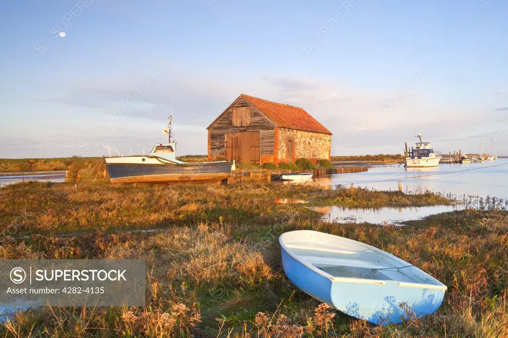 England, Norfolk, Thornham. Boats and the old coal shed at first light at Thornham Harbour during a high tide on the North Norfolk Coast.