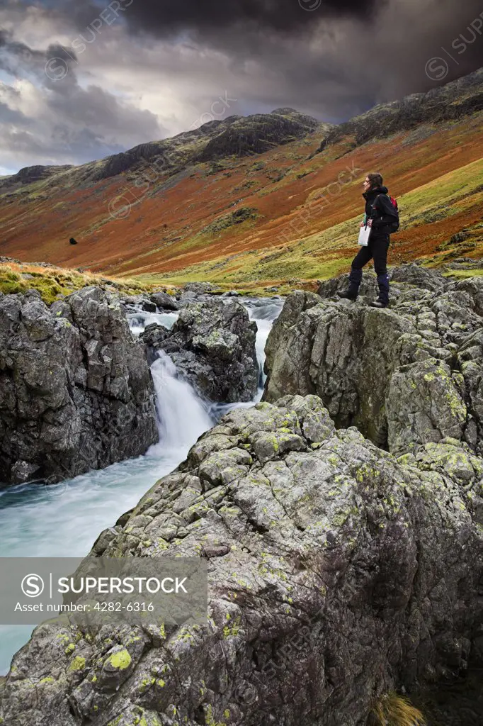 England, Cumbria, Brotherilkeld. Young woman standing on boulders next to the River Esk. The River Esk begins in Great Moss below Scafell Pike in the English Lake District and flows down to the sea at Ravenglass.