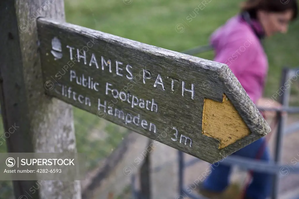 England, Oxfordshire. A walker with her dog passing through a gate beneath a signpost on the Thames Path showing the direction and mileage to Clifton Hampden.